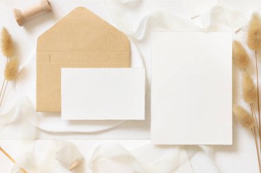 Kıvrılmış ipek kurdelelerin yanında boş kartlar ve zarf ve kurutulmuş tavşanın kuyruk çimlerinin üst görünümü beyaz, maket. Pastel romantik düz yatak. Parti tasarımı ya da düğün kırtasiyesi için bohem ya da doğa kartlarına yakın