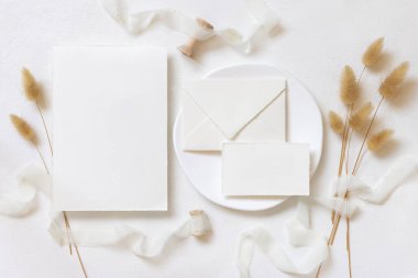 Kıvrılmış ipek kurdelelerin yanında boş kartlar ve zarf ve kurutulmuş tavşanın kuyruk çimlerinin üst görünümü beyaz, maket. Pastel romantik düz yatak. Parti tasarımı ya da düğün kırtasiyesi için bohem ya da doğa kartlarına yakın
