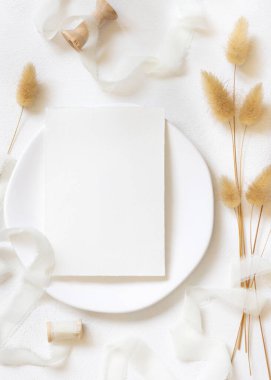 Kıvrımlı ipek kurdelelerin yanındaki boş kart ve kurutulmuş tavşanın kuyruğundaki çim beyaz, maket. Boho Pastel Romantik Flatlay. Parti tasarımı ya da düğün kırtasiyesi için bohem ya da doğa kartlarına yakın