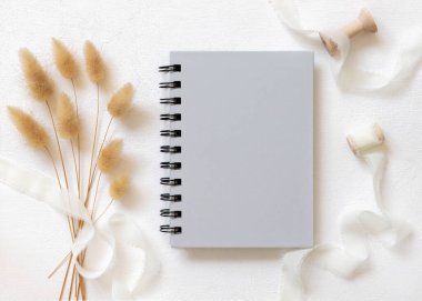 Kıvrımlı ipek kurdelelerin yanında gri, boş, sarmal bir defter ve kurutulmuş tavşanın kuyruk çimlerini beyazın üstünde, maket. Pastel romantizm sahnesi ve ciltli ders kitabı. Bohem ya da doğaya yakın kız gibi yatıyordu.