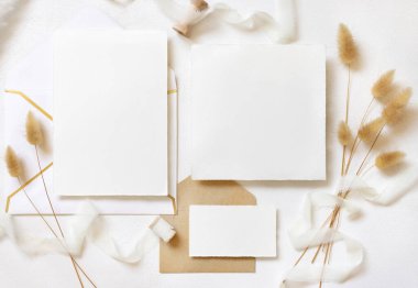 Bukleli ipek kurdelelerin yanında boş kartlar ve zarflar ve kurutulmuş tavşanın kuyruk çimlerini beyazın üstünde, maket. Pastel romantik düz yatak. Parti tasarımı ya da düğün kırtasiyesi için bohem ya da doğa kartlarına yakın