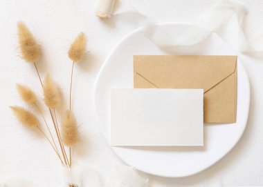 Beyaz plaka üzerine boş kart ve zarf kıvrılmış ipek kurdeleler ve kurutulmuş tavşanın kuyruk çimen üst görünümü, maket. Pastel romantik düz yatak. Parti tasarımı ya da düğün kırtasiyesi için bohem ya da doğa kartlarına yakın