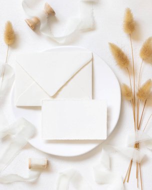 Beyaz plaka üzerine boş kart ve zarf kıvrılmış ipek kurdeleler ve kurutulmuş tavşanın kuyruk çimen üst görünümü, maket. Pastel romantik düz yatak. Parti tasarımı ya da düğün kırtasiyesi için bohem ya da doğa kartlarına yakın