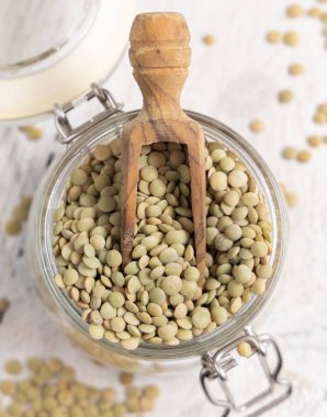 Beyaz masa manzaralı ahşap bir kepçe ile kurutulmuş yeşil mercimek dolu cam kavanoz. Sağlıklı vejetaryen yiyecekler, bitkisel protein kaynakları. Geleneksel Akdeniz çiğ malzemesi