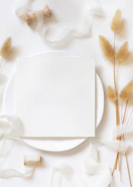 Kıvrımlı ipek kurdelelerin yanındaki boş kare kart ve kurutulmuş tavşanın kuyruk çimlerini beyaz, model. Boho Pastel Romantik Flatlay. Parti tasarımı ya da düğün kırtasiyesi için bohem ya da doğa kartlarına yakın