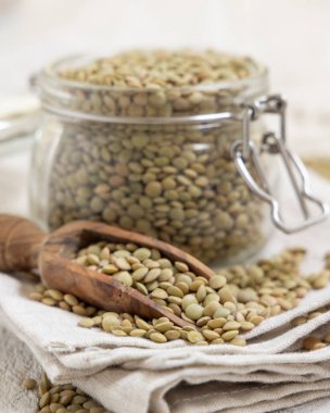 Kurumuş yeşil mercimeklerle dolu açık cam kavanoz ve yakın plan beyaz masadaki ahşap bir kepçe. Sağlıklı vejetaryen yiyecekler, bitkisel protein kaynakları. Geleneksel Akdeniz çiğ malzemesi