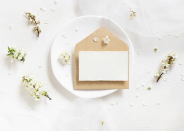 Tülün yanındaki tabakta boş kart ve kraft kağıt zarf ve yaban erikleri beyaz çiçekler üst manzara, maket. Bahar partisi tasarımı için Pastel yatay kart, anneler günü veya romantik düğün kırtasiyesi