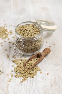 Kurumuş yeşil mercimeklerle dolu açık cam kavanoz ve beyaz masa üzerinde ahşap bir kepçe. Sağlıklı vejetaryen yiyecekler, bitkisel protein kaynakları. Geleneksel Akdeniz çiğ malzemesi