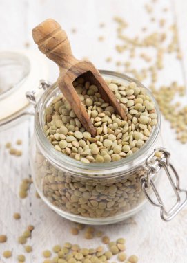 Kurumuş yeşil mercimeklerle dolu açık cam kavanoz ve beyaz masa üzerinde ahşap bir kepçe. Sağlıklı vejetaryen yiyecekler, bitkisel protein kaynakları. Geleneksel Akdeniz çiğ malzemesi