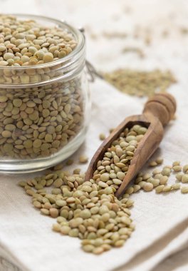 Kurumuş yeşil mercimeklerle dolu açık cam kavanoz ve yakın plan beyaz masadaki ahşap bir kepçe. Sağlıklı vejetaryen yiyecekler, bitkisel protein kaynakları. Geleneksel Akdeniz çiğ malzemesi