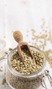 Kurumuş yeşil mercimeklerle dolu açık cam kavanoz ve beyaz masa üzerinde ahşap bir kepçe. Sağlıklı vejetaryen yiyecekler, bitkisel protein kaynakları. Geleneksel Akdeniz çiğ malzemesi