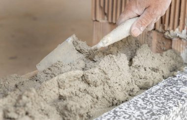 Zanaatkar eli tozla kaplı. Duvara beton ya da alçı yapıştırıyor. Yakın plan. Mala ve kovayı elinde tutan işçiler. İnşaat veya yenileme süreci ayrıntıları