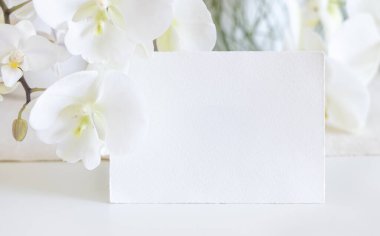 Beyaz orkide çiçeklerinin yanındaki boş kart. Masaya yakın, maket. Egzotik düğün, Sevgililer Günü ya da Anneler Günü kırtasiyesi ve tropikal parti tasarımı için yatay kartlı Pastel romantik sahne