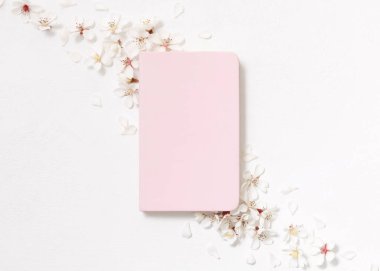 Beyaz kiraz çiçeklerinin yanındaki açık pembe defter beyaz masanın üstündeki üst görünüm, kopyalama alanı, maket. Okul ya da mandıra için Spiral ciltli ders kitabı, bahar havası