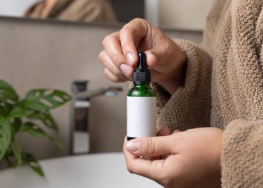 Doğal manikürlü eller yeşil kozmetik şişesi tutuyor, etiket modeli. Banyoda üzerinde bitki ve musluk olan kahverengi bornozlu bir kadın. 