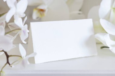 Beyaz orkide çiçeklerinin yanında katlanmış boş yer kartı. Masaya yakın, maket. Egzotik düğün, Sevgililer Günü ya da Anneler Günü kırtasiyesi ve tropikal parti tasarımı için yatay kartlı Pastel romantik sahne