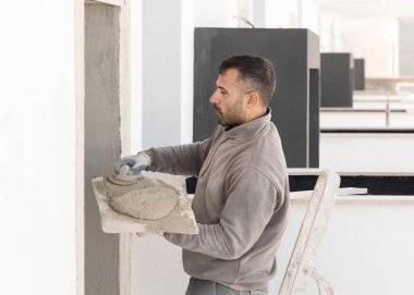 Koyu renk saçlı genç bir zanaatkar kapı eşiğine metal yüzgeçli sıva serpiştiriyor. İnşaat ve yenileme sürecinde kirli eldivenli ve gri iş elbiseli adamlar