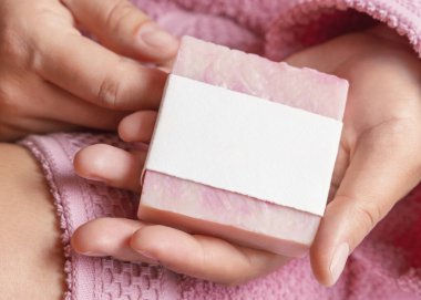 Doğal manikürlü kadın elleri boş etiketli sabun kalıbı, ambalaj modeli. Banyoda açık pembe bornoz giyen bir kadın. Hijyen kavramı