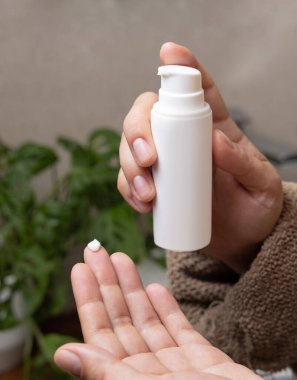 Kahverengi bornozlu bir kadının elinde beyaz, boş bir pompa şişesi, kozmetik model. Yeşil bitki ve leğene karşı banyoda günlük vücut bakımı rutini. Cilt bakım ürünleri