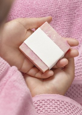 Doğal manikürlü kadın elleri pembe bornozun üzerinde boş etiketli sabun kalıbı, ambalaj modeli. Doğal manikürlü genç bir kız. Hijyen kavramı