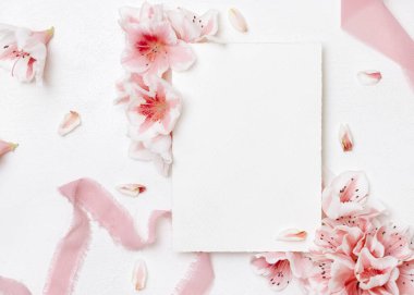 Pembe açelya çiçeklerinin ve ipek kurdelelerin yanındaki boş kağıt kart. Üst manzara, fotokopi alanı, maket. İlkbahar romantik düğün kırtasiyesi ya da parti tasarımı için dikey kart, Anneler Günü ya da doğum günü havası