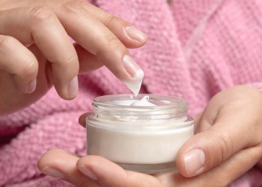 Pembe bornozlu, kremalı, parmaklı ve elinde kozmetik kavanozu olan genç bir kadının yakın plan manikürlü fotoğrafını çek. Yüz bakımı için kız organik kozmetik ürünleri