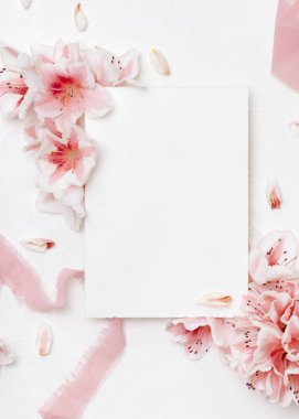 Pembe açelya çiçeklerinin ve ipek kurdelelerin yanındaki boş kağıt kart. Üst manzara, fotokopi alanı, maket. İlkbahar romantik düğün kırtasiyesi ya da parti tasarımı için dikey kart, Anneler Günü ya da doğum günü havası