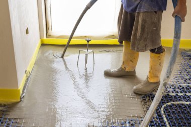 Sarı lastik çizmeli bir adam yeni binadaki ısıtma tesisatına sıvı beton döküyor, plastik borular ve boru hatları siyah yalıtım çarşafına bağlanıyor. Modern ısıtma sistemi