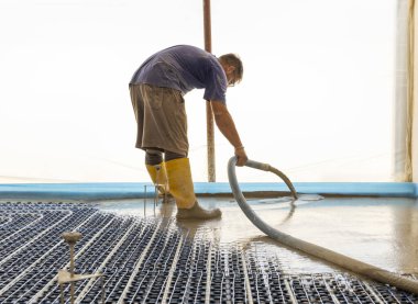 Sarı lastik çizmeli bir adam yeni binadaki ısıtma tesisatına sıvı beton döküyor, plastik borular ve boru hatları siyah yalıtım çarşafına bağlanıyor. Modern ısıtma sistemi