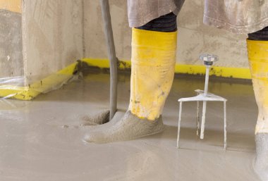 Sarı lastik çizme giyen bir adam yeni binadaki ısıtma tesisatının üzerine sıvı beton döküyor. Yeni ev inşaatında modern ısıtma sistemi