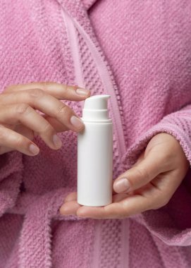 Pembe bornozlu genç bir kadının elinde beyaz bir pompa krem tüpü, kız makyajlı bir model. Kızın doğal manikürü var. Her gün yüz bakımı ya da bakım rutini, spa prosedürleri