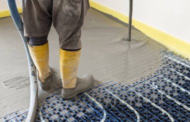 Sarı lastik çizmeli bir adam yeni binadaki ısıtma tesisatına sıvı beton döküyor, plastik borular ve boru hatları siyah yalıtım çarşafına bağlanıyor. Modern ısıtma sistemi