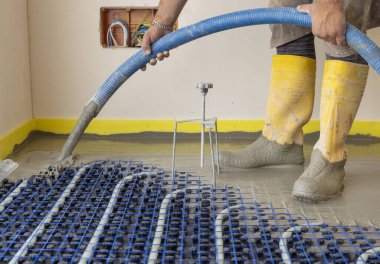Sarı lastik çizmeli bir adam yeni binadaki ısıtma tesisatına sıvı beton döküyor, plastik borular ve boru hatları siyah yalıtım çarşafına bağlanıyor. Modern ısıtma sistemi