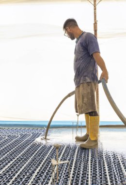 Sarı lastik çizmeli bir adam yeni binadaki ısıtma tesisatına sıvı beton döküyor, plastik borular ve boru hatları siyah yalıtım çarşafına bağlanıyor. Modern ısıtma sistemi