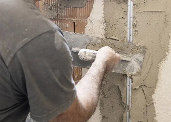 Zanaatkar, evin duvarını kaplamak için şamandıra ve çimento kullanıyor. İnşaat ve yenileme çalışmaları sırasında eldivenli ve gri elbiseli adamlar