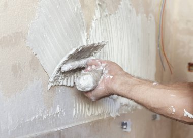 Zanaatkar fayans yapıştırıcısını fayanssız bir duvar yüzeyine çentik malayla yayar, eller yakın çekim yapar. Yeni bina inşaatı veya yenilenmesi.