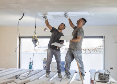 İki tozlu zanaatkar, iskelede ayakta duran bir odanın tavanındaki sıvaları kaymak için mala ve şahin kullanır. İnşaat veya yenileme sürecinde gri iş elbiseli erkekler