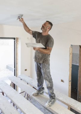 Tozlu bir zanaatkar, iskelede ayakta duran bir odanın tavanının kaymağını sıyırmak için mala ve şahin kullanır. İnşaat sırasında ya da bir evin tadilatı sırasında gri iş elbiseli adamlar