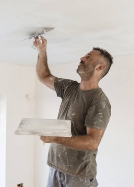 Tozlu bir zanaatkar, iskelede ayakta duran bir odanın tavanının kaymağını sıyırmak için mala ve şahin kullanır. İnşaat sırasında ya da bir evin tadilatı sırasında gri iş elbiseli adamlar