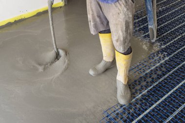 Sarı lastik çizmeli bir adam yeni binadaki ısıtma tesisatına sıvı beton döküyor, plastik borular ve boru hatları siyah yalıtım çarşafına bağlanıyor. Modern ısıtma sistemi