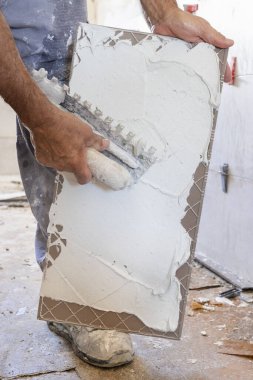 Zanaatkar fayans yapıştırıcısını fayans sırtında çentik olan bir malayla yayar, eller yakın çekimde. Yeni bina inşaatı veya tadilatı, DIY ev tadilatı