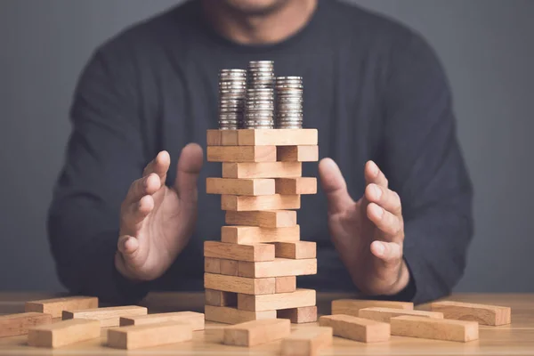 Risks in business or financial concept. Idea to prevent risk in business. Business man using hand to protection coin stacked on tower wooden block game and prevent falling down. Studio shot.