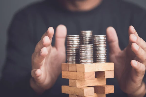 Risks in business or financial concept. Idea to prevent risk in business. Business man using hand to protection coin stacked on tower wooden block game and prevent falling down. Studio shot.
