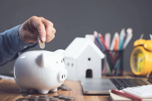 House purchase and money saving idea, Home cost calculate concept. Man adding coin to white piggy bank on office desk to plan for house financial. Residence buying planning in the future. Studio shot.