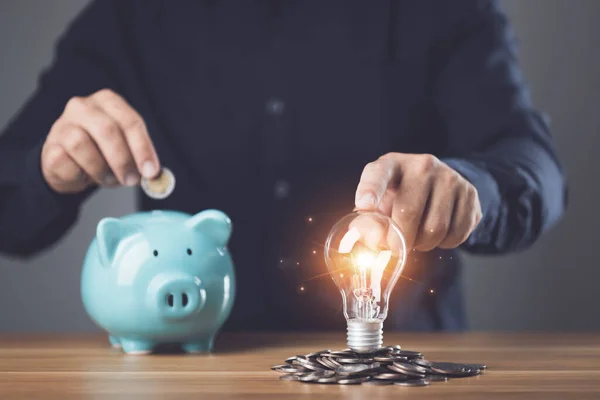 Business, Financial and money planning, Clever thinking about money saving for future and retirement, Man holding light bulb with coin pile and adding coin to piggy bank on the table. Studio shot.