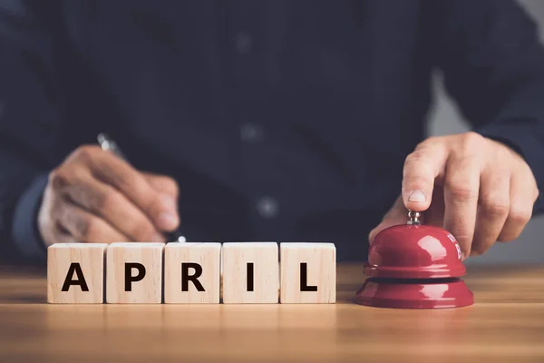 Notification new month has begun, Close up hand ringing the red call bell or service bell ring and word APRIL arranged on wooden desk. For hotel or restaurant advertising concept. Studio shot.