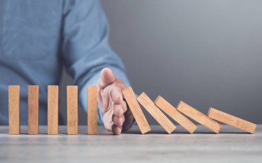 İşadamı, domino etkisi ve risk yönetimini temsil eden tahta blokların düşmesini engellemek için elini kullanıyor. Sorunların artmasını önlemek ve risklerin etkili bir şekilde yönetilmesini sembolize eder