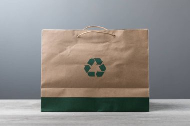 Geri dönüşüm sembollü kahverengi kese kağıdı masaya konur. Görüntü, sürdürülebilirlik ve çevre dostu uygulamalar kavramını vurgulayarak, materyallerin yeniden kullanılması ve geri dönüştürülmesini teşvik etmektedir..