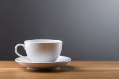 Beyaz kahve fincanı, koyu gri arka planda, ahşap bir masanın üzerinde duruyor. Görüntü sıcaklığı, sadeliği ve bir fincan taze kahvenin keyfini çıkartıyor.