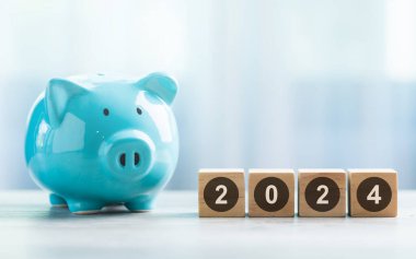 2024 is written on a wooden block beside a A blue piggy bank on a wooden table against a bright white background. Money planning and Financial planning, savings, and wise money management. clipart